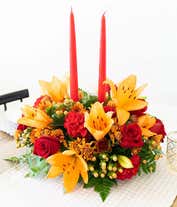 Alternative image 1 for A stunning floral centerpiece featuring red roses, red carnations, orange lilies, peach berries, and bronze cushion poms, complemented by two orange taper candles.