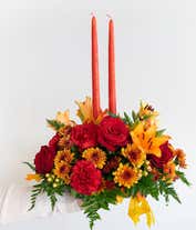 Alternative image 2 for A stunning floral centerpiece featuring red roses, red carnations, orange lilies, peach berries, and bronze cushion poms, complemented by two orange taper candles.