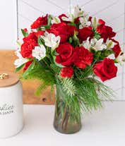 Alternative image 1 for A festive arrangement with red roses, red carnations, red mini carnations, white alstroemeria, and evergreens, beautifully displayed in a glass vase.