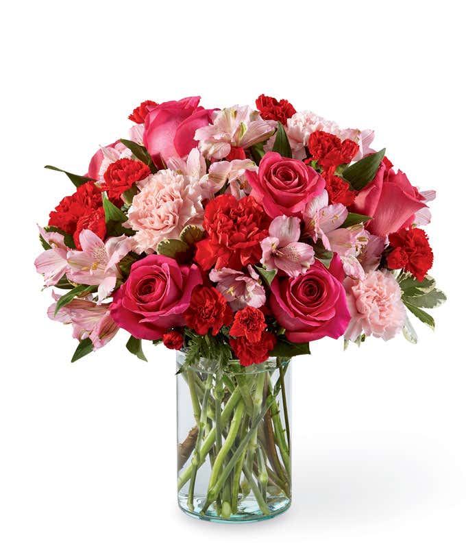 Pink roses, carnations, alstroemeria, red carnations, and fresh floral greens in a clear cylinder vase