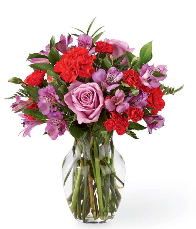 Lavender roses, purple alstroemeria, red carnations, and fresh floral greens in a clear glass vase