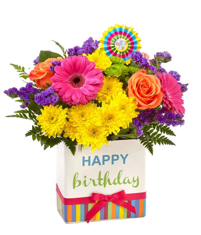 Orange roses, pink gerbera daisies, yellow mums, purple filler, and fresh floral greens with a birthday pick, in a cube vase that says happy birthday