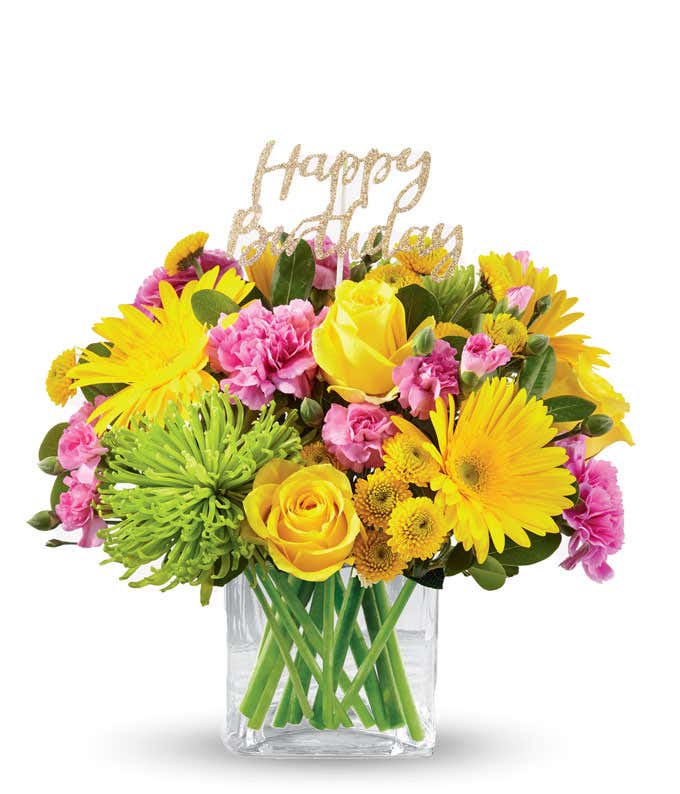 Yellow daisies and roes, pink carns, and green mums with a gold happy birthday pick, in a clear cube vase
