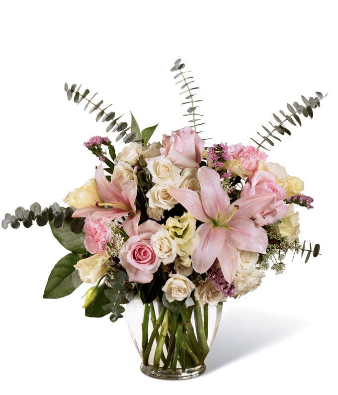 Light pink lilies, and white roses are arranged with mixed greens into a clear glass vase