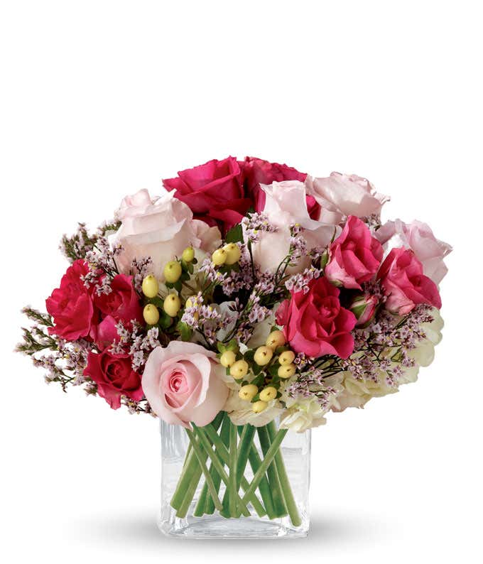 Short arrangement of light and dark pink roses, with white hydrangea, and huckleberry in a cube vase