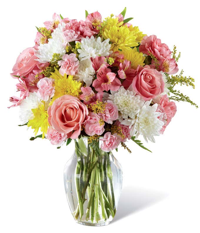 Full and round arrangement of pink roses, carns, and alstroemeria, mixed with white and yellow poms in a clear glass vase