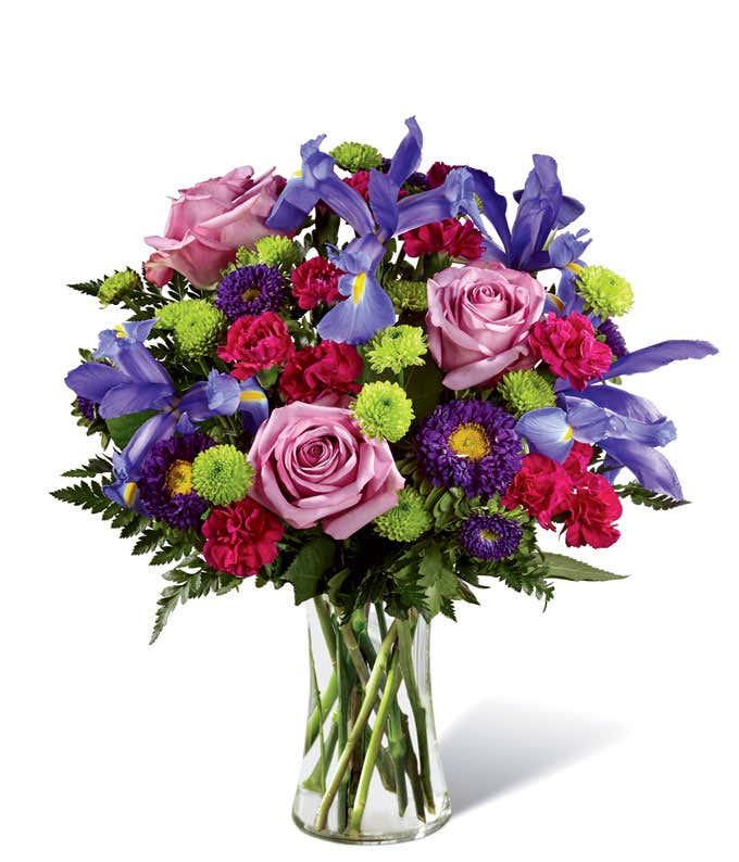 Tall arrangement of purple roses, iris, and green and purple asters, all in a cylinder vase