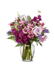 A bouquet of lavender roses, purple alstroemeria and statice, with fresh floral greens, in a clear glass vase 