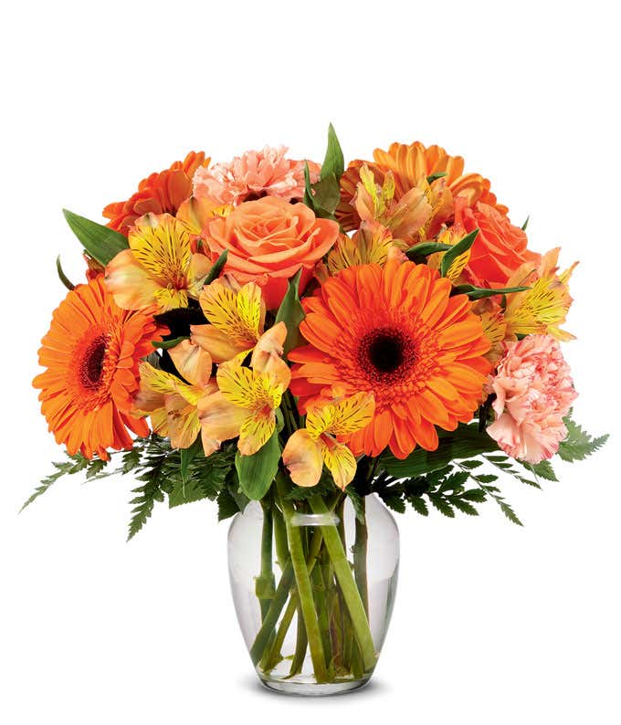 Orange gerbera daisies, roses, alstroemeria, and carnations, with fresh floral greens in a clear glass vase