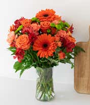 Alternative image 4 for A bold floral arrangement featuring orange roses, orange gerbera daisies, red alstroemeria, and green button poms, all beautifully arranged in a glass vase. This vibrant mix offers a striking and energetic display.