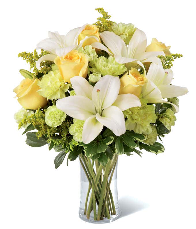 White lilies, yellow roses, and green carnations, arranged in a tall clear cylinder vase