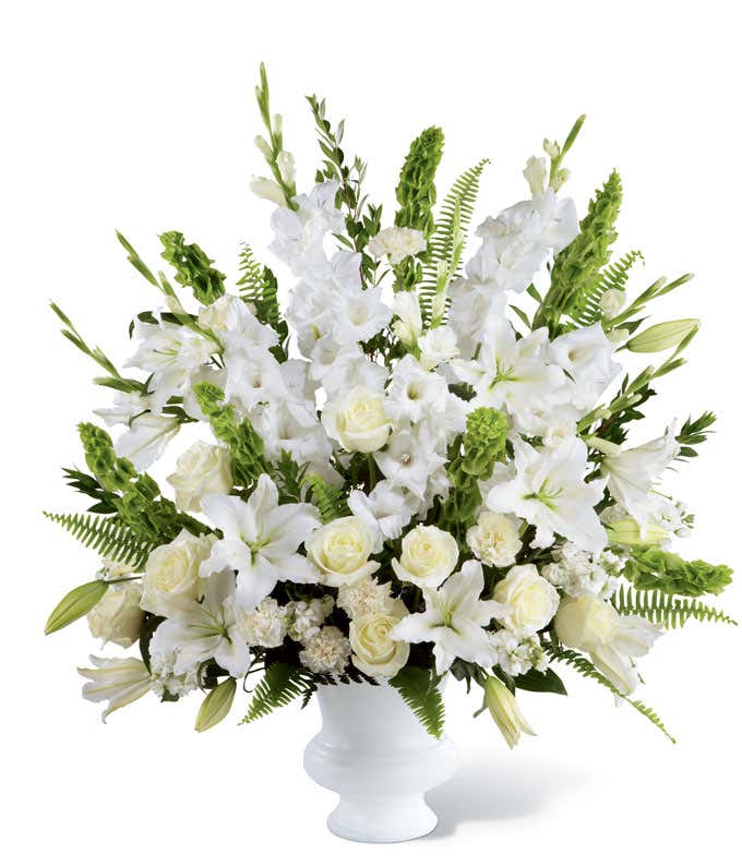 A standing basket of white roses, lilies, gladiolus, bells of Ireland, and fresh greens, in a white plastic urn.