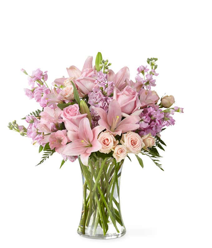 A bouquet of pink lilies, roses, stock and alstroemeria, with fresh floral greens in a tall cylinder vase