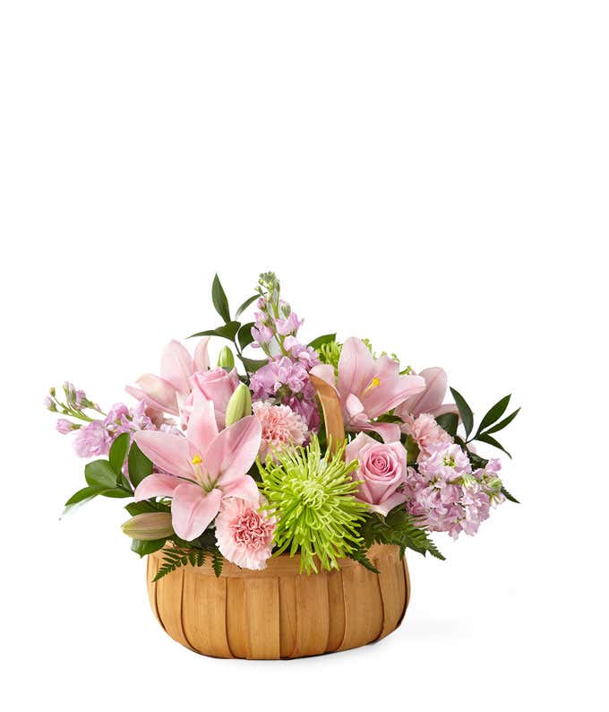Pink lilies, roes, and carnations, with green poms and fresh floral greens arranged into a wooden basket