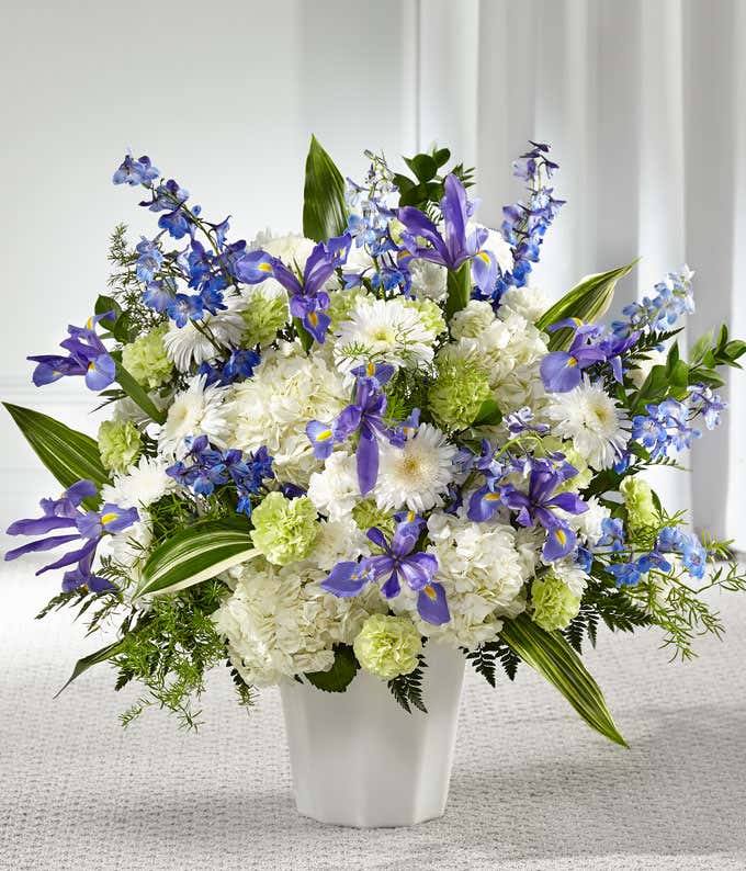 A standing basket of white hydrangeas, blue delphinium, iris, white poms, green carnations, and fresh floral greens in a white plastic urn