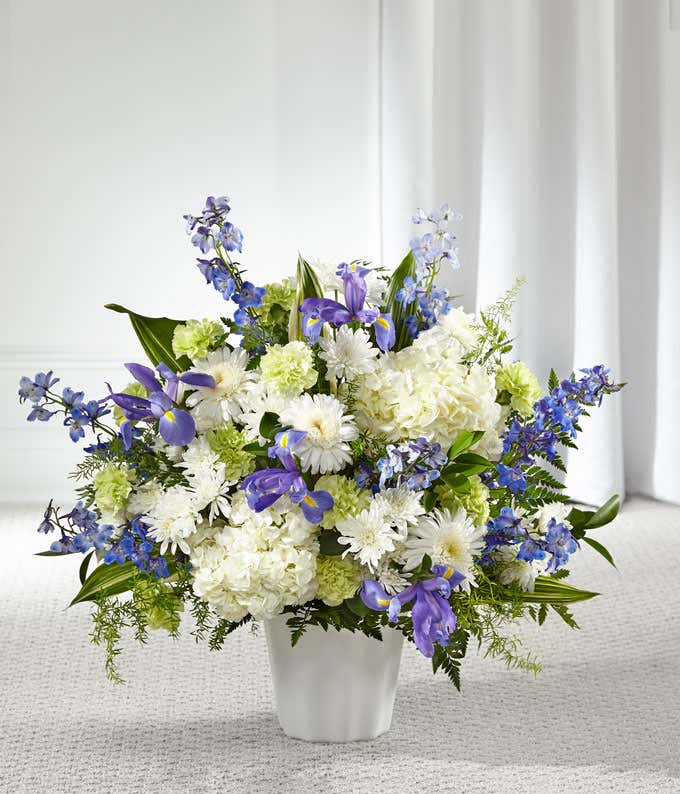A standing basket of white hydrangeas, blue delphinium, iris, white poms, green carnations, and fresh floral greens in a white plastic urn