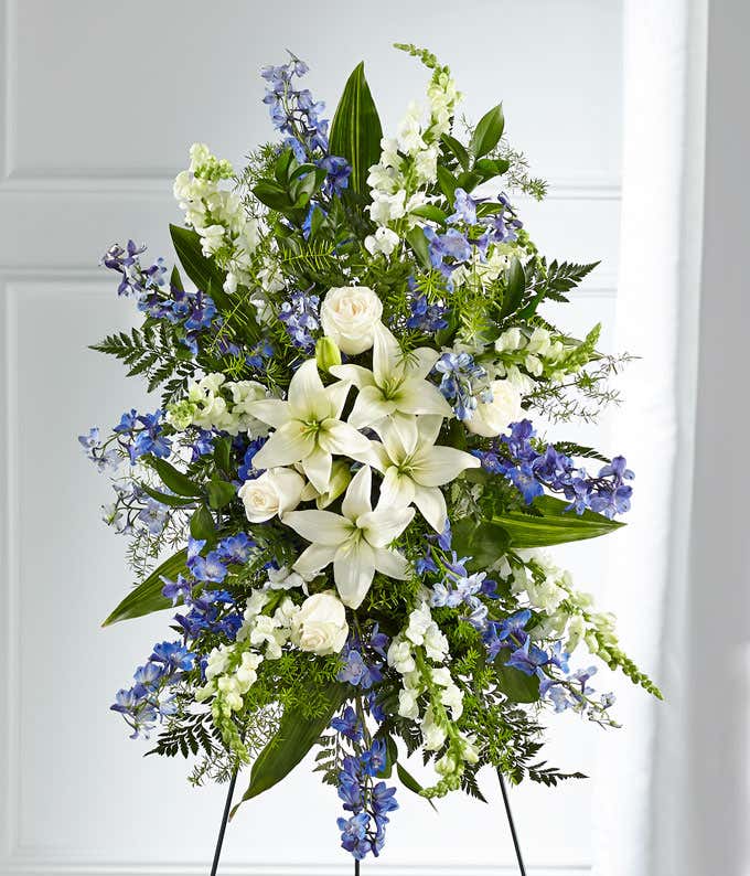 A standing spray of white lilies, roses, and snapdragons, with blue delphinium and fresh floral greens