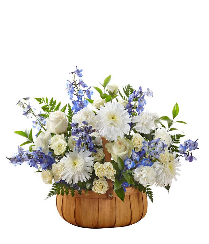 White roses, poms, and carnations, with blue delphinium and fresh floral greens, arranged in a wooden basket