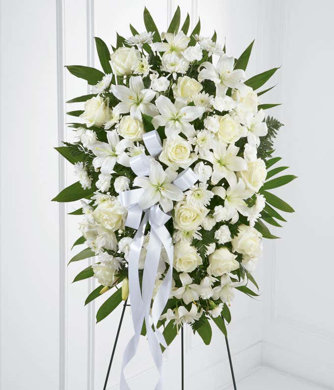 A standing spray made up of white lilies, roses, mums, and carnations, with fresh floral greens and a white ribbon