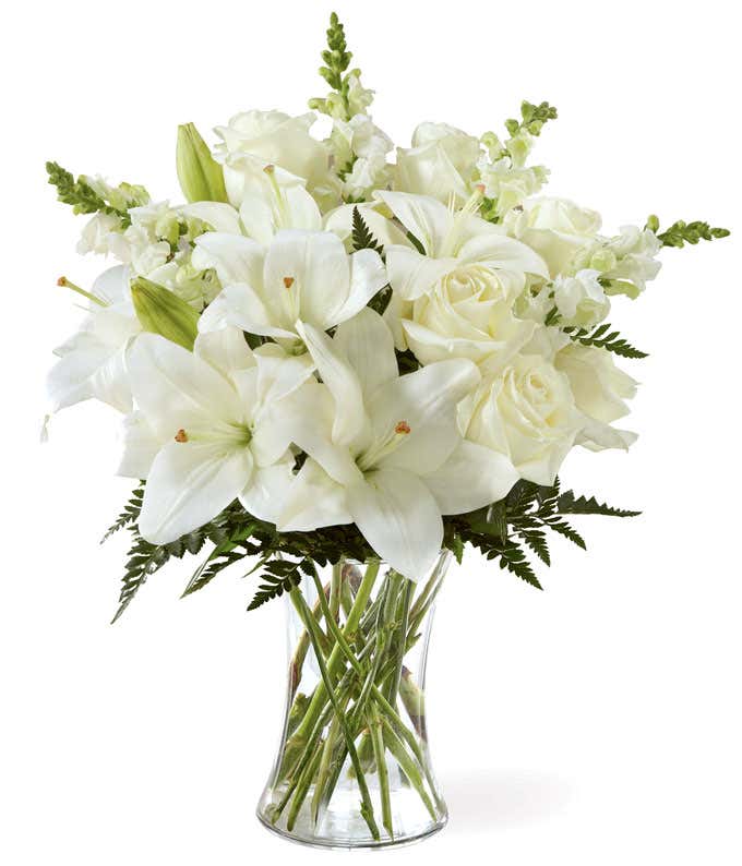 A bouquet of white lilies, roses, and snapdragons, with fresh floral greens, in a tall clear cylinder vase