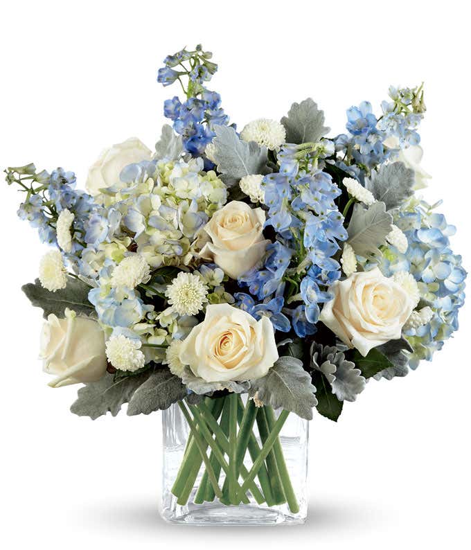 White roses, blue hydrangea, blue delphinium, and fresh greens arranged into a short cube vase