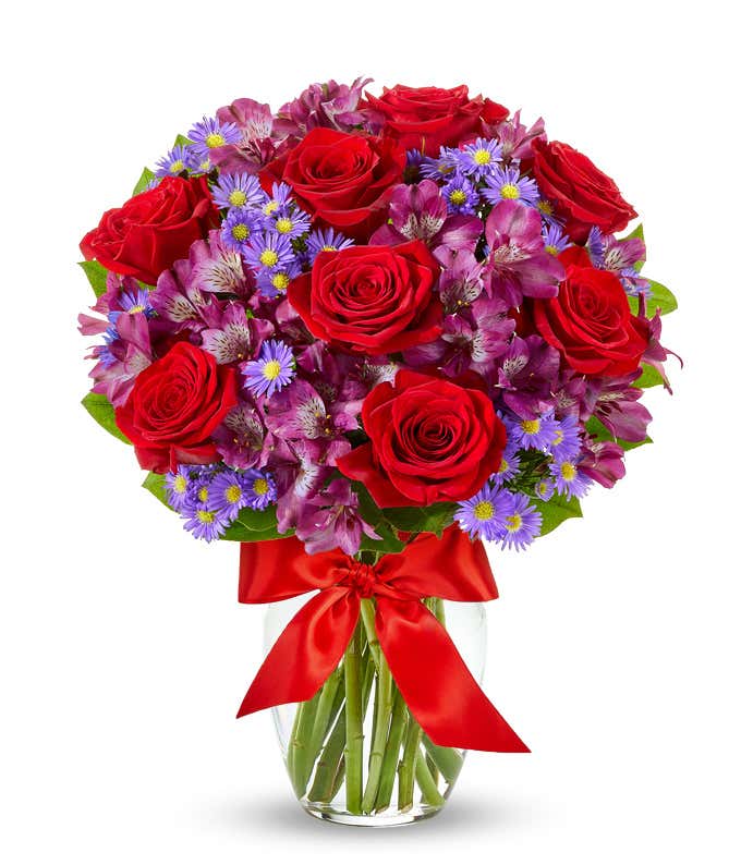 A bouquet with red roses, purple alstroemeria, and purple daisy poms in a clear vase tied with a red ribbon.