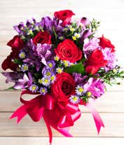 Alternative image 1 for A bouquet with red roses, purple alstroemeria, and purple daisy poms in a clear vase tied with a red ribbon.