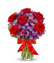 A bouquet with red roses, purple alstroemeria, and purple daisy poms in a clear vase tied with a red ribbon.