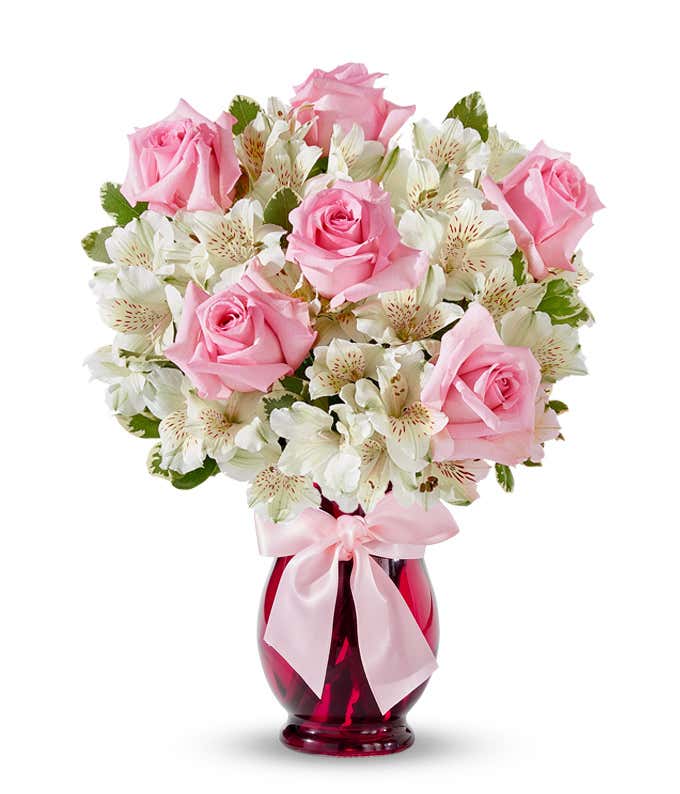 A bouquet with pink roses and white alstroemeria in a red vase, tied with a pink ribbon.