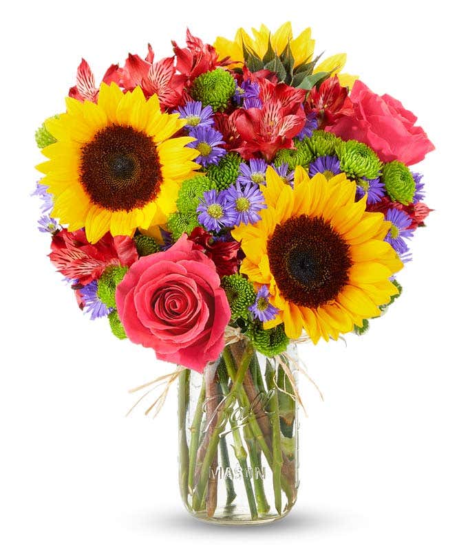 A vibrant bouquet in a mason jar featuring large yellow sunflowers, pink roses, red alstroemeria, purple poms, and green chrysanthemums. The jar is wrapped with a simple twine bow, showcasing the fresh green stems inside.