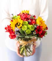 Alternative image 1 for A vibrant bouquet in a mason jar featuring large yellow sunflowers, pink roses, red alstroemeria, purple poms, and green chrysanthemums. The jar is wrapped with a simple twine bow, showcasing the fresh green stems inside.
