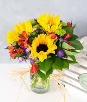 Alternative image 2 for A vibrant bouquet in a mason jar featuring large yellow sunflowers, pink roses, red alstroemeria, purple poms, and green chrysanthemums. The jar is wrapped with a simple twine bow, showcasing the fresh green stems inside.