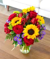 Alternative image 3 for A vibrant bouquet in a mason jar featuring large yellow sunflowers, pink roses, red alstroemeria, purple poms, and green chrysanthemums. The jar is wrapped with a simple twine bow, showcasing the fresh green stems inside.