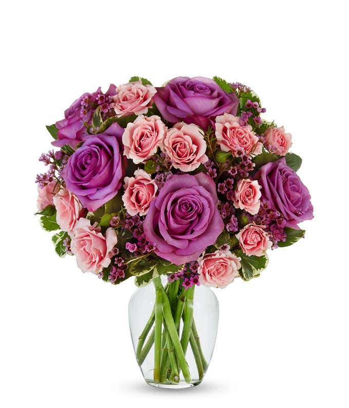  A bouquet in a clear vase featuring large purple roses, light pink roses, and small pink waxflowers. The arrangement is lush and rounded, with green leaves adding contrast and depth, and the fresh green stems visible through the vase.