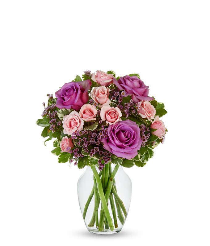  A bouquet in a clear vase featuring large purple roses, light pink roses, and small pink waxflowers. The arrangement is lush and rounded, with green leaves adding contrast and depth, and the fresh green stems visible through the vase.