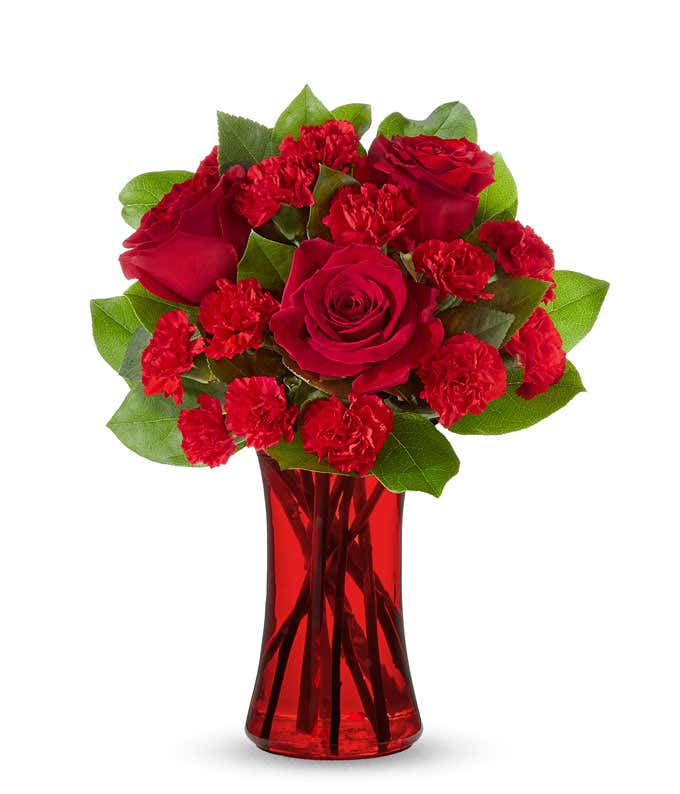 A striking arrangement featuring red roses, red mini carnations, and salal, elegantly presented in a red gathering vase. This bold and vibrant display is perfect for adding a touch of elegance and passion to any occasion.