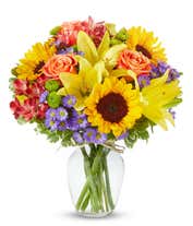 A radiant floral arrangement featuring sunflowers, yellow lilies, orange roses, alstroemeria, Monte Casino, and green poms, all beautifully arranged in a clear glass vase. This vibrant mix creates a warm and inviting display.