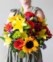 Alternative image 3 for A radiant floral arrangement featuring sunflowers, yellow lilies, orange roses, alstroemeria, Monte Casino, and green poms, all beautifully arranged in a clear glass vase. This vibrant mix creates a warm and inviting display.