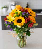 Alternative image 4 for A radiant floral arrangement featuring sunflowers, yellow lilies, orange roses, alstroemeria, Monte Casino, and green poms, all beautifully arranged in a clear glass vase. This vibrant mix creates a warm and inviting display.