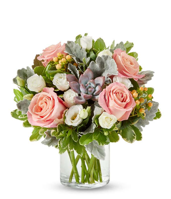 A bouquet in a clear vase with pink roses, white lisianthus, a succulent centerpiece, and accents of green leaves and berries.