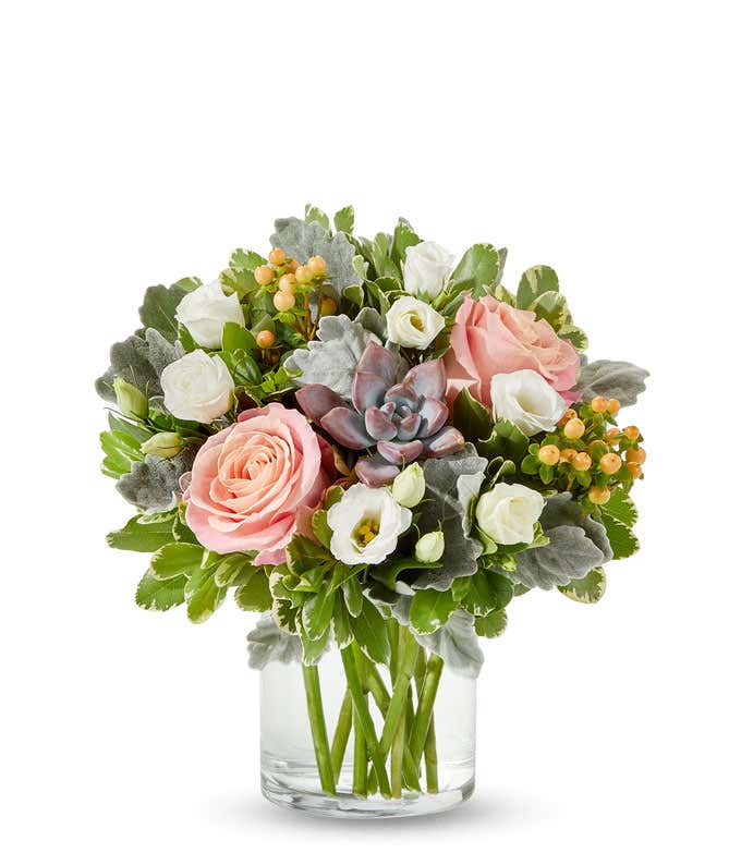 A bouquet in a clear vase with pink roses, white lisianthus, a succulent centerpiece, and accents of green leaves and berries.