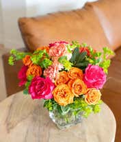 Alternative image 1 for A lively floral arrangement featuring hot pink roses, orange spray roses, orange carnations, bupleurum, lily grass, and pittosporum, all beautifully arranged in a clear glass cube vase. This vibrant mix creates a stunning and energetic display.