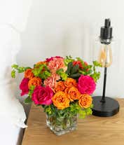Alternative image 2 for A lively floral arrangement featuring hot pink roses, orange spray roses, orange carnations, bupleurum, lily grass, and pittosporum, all beautifully arranged in a clear glass cube vase. This vibrant mix creates a stunning and energetic display.
