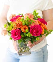 Alternative image 3 for A lively floral arrangement featuring hot pink roses, orange spray roses, orange carnations, bupleurum, lily grass, and pittosporum, all beautifully arranged in a clear glass cube vase. This vibrant mix creates a stunning and energetic display.