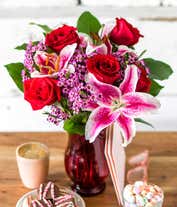 Alternative image 1 for A striking arrangement featuring red roses, stargazer lilies, waxflower, and seasonal greens, elegantly presented in a red cylinder vase. This bold and vibrant display is perfect for adding a touch of elegance and passion to any occasion.