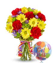 A cheerful floral arrangement featuring red carnations, yellow daisies, purple Monte Casino, and green button poms in a vase decorated with rainbow ribbons, accompanied by a 'Get Well' mylar balloon.