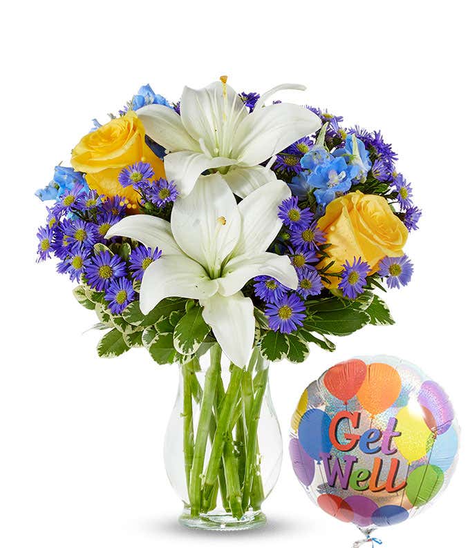 A floral arrangement featuring white Asiatic lilies, yellow roses, blue delphinium, and purple Monte Casino in a keepsake glass vase, accompanied by a 'Get Well' mylar balloon.