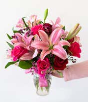 Alternative image 2 for A romantic arrangement featuring red and pink roses, pink lilies, purple waxflowers, and alstroemeria, beautifully arranged in a clear vase with a bow. Includes a keepsake teddy bear and a box of chocolate for an extra touch of love.