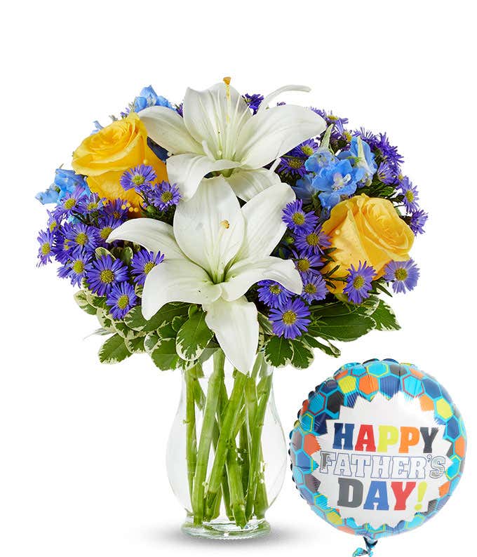 White lilies, yellow roses, and tall blue delphinium poke out from a clear vase. Theyr'e accented by variegated green leaves and small purple daisy flowers. Attached is a Happy Father's Day Mylar balloon on a string.