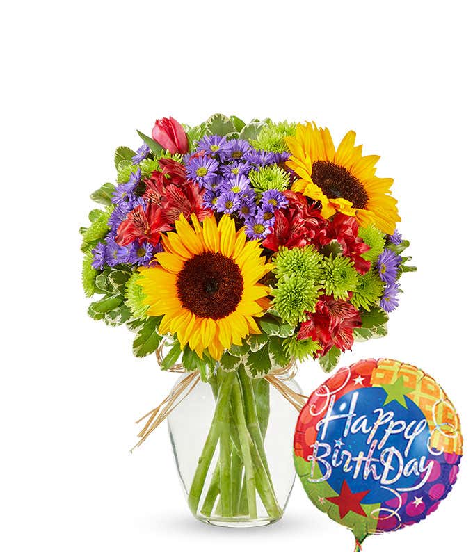 a colorful bouquet in a clear vase, featuring bright yellow sunflowers, red tulips, purple asters, and lush green foliage. The arrangement is tied with a rustic twine bow. Next to the flowers there's a 'Happy Birthday' mylar balloon.